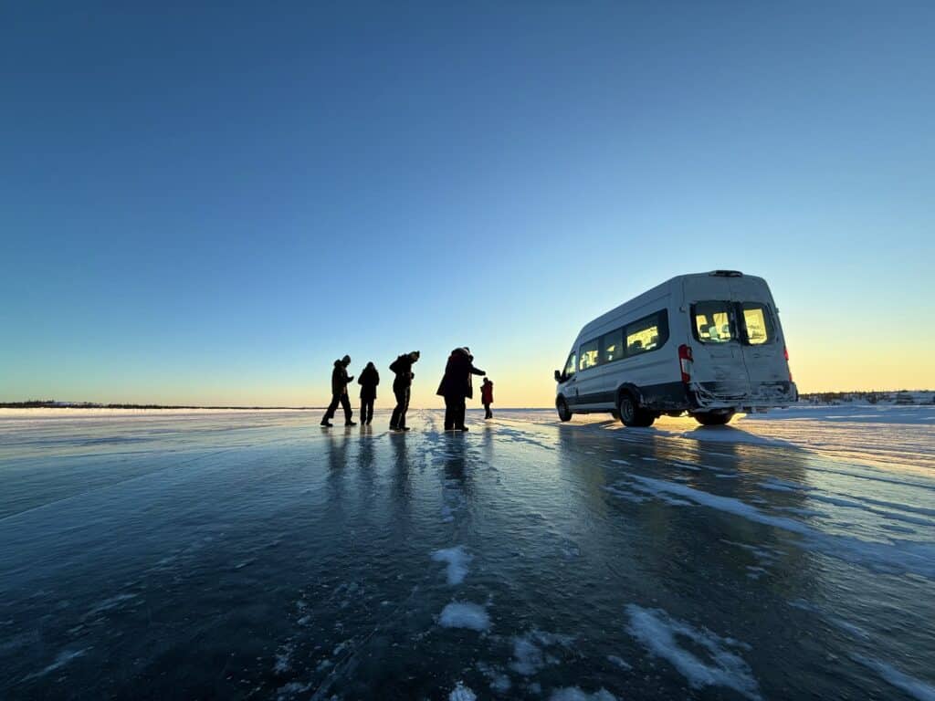 group of people stand on ice road beside tour van with the setting sun behind them