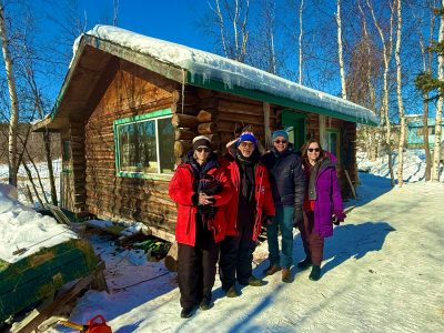 Group of people stand outside of the trapper cabin at dogsled